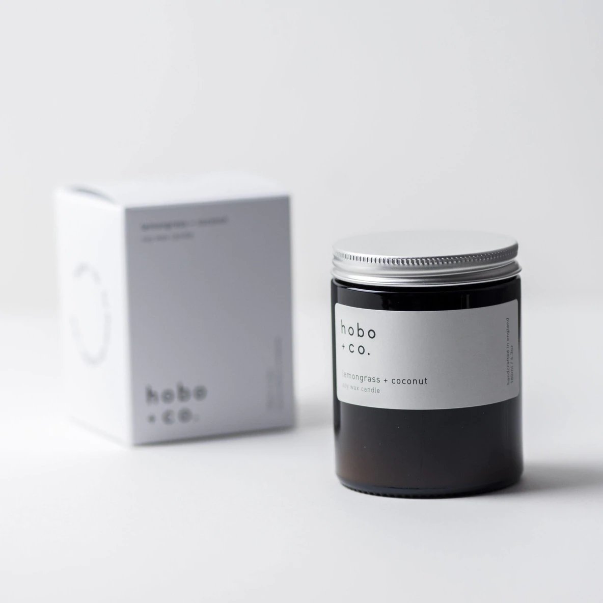 3CBD172F-6A3D-4163-93A9-4457F8B30709 Eine Duftkerze von hobo + co. mit Zitronengras und Kokosnuss, neben der Verpackung auf einem weißen Hintergrund.