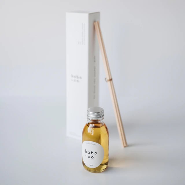 B2FA7882-E2A9-401D-9A9C-2EF7572F1F83 Kleine Flasche mit Duftöl von hobo + co. und Holzstäbchen vor einer weißen Verpackung auf einem weißen Hintergrund.