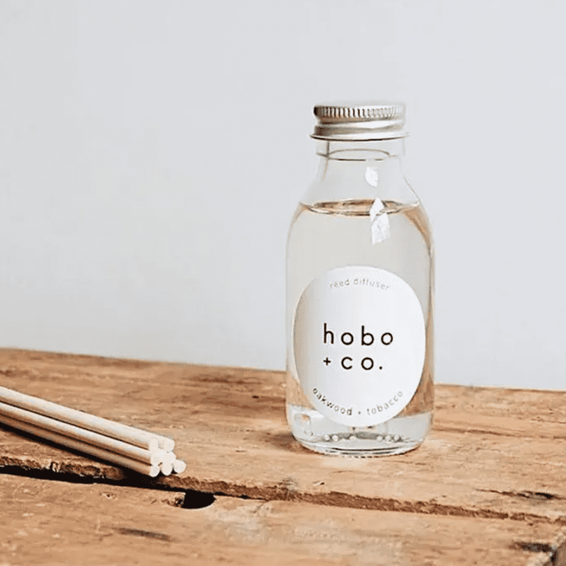 hobo-co-oakwood-tobacco-reed-diffuser_optimized Hobo + Co. Oakwood Tobacco Reed Diffuser auf einem Holztisch mit Duftstäbchen.