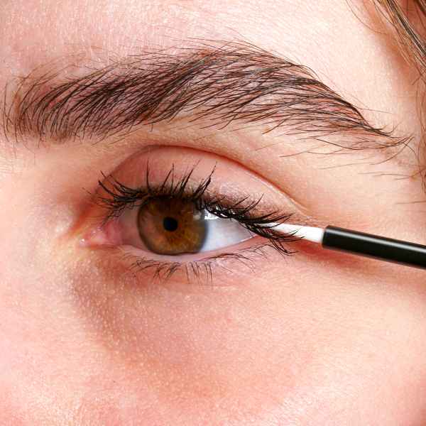 Nahaufnahme eines Auges mit brauner Iris, auf das ein Pinsel Wimpernserum aufträgt.
