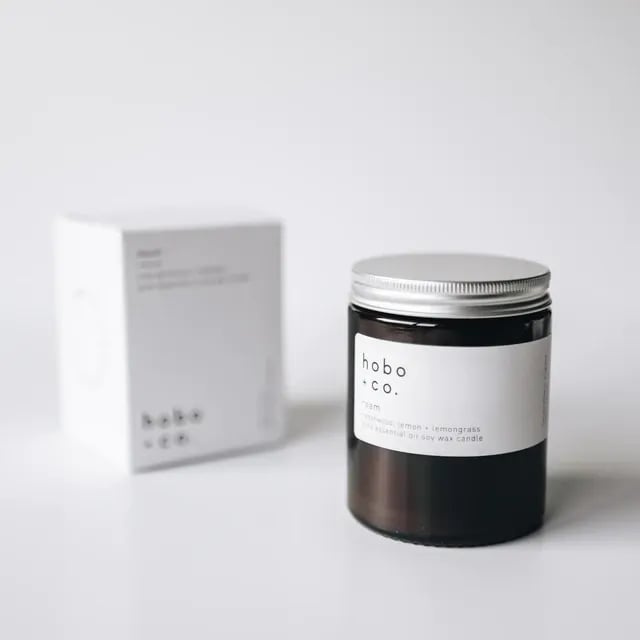 Eine schwarze Duftkerze von hobo + co. mit der Aufschrift 'Cedarwood, Lemon + Lemongrass' steht vor einer weißen Verpackung.